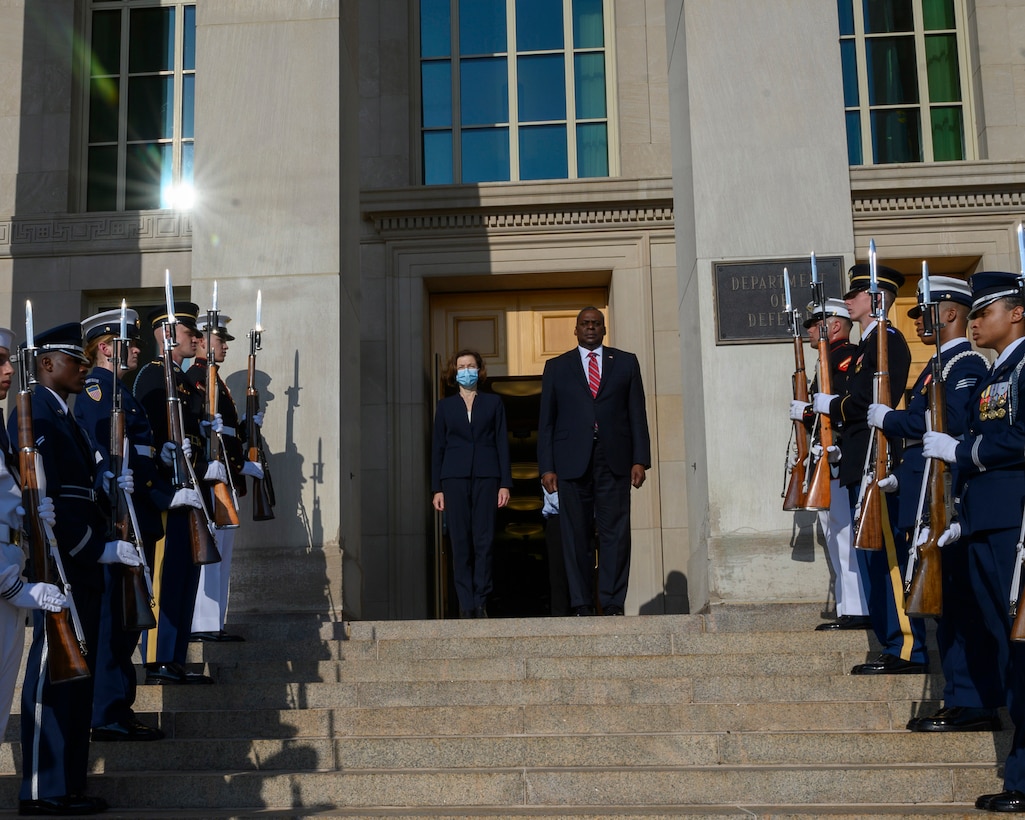 A man and a woman stand at the top of a series of steps leading up to an office building. Military personnel line the steps.