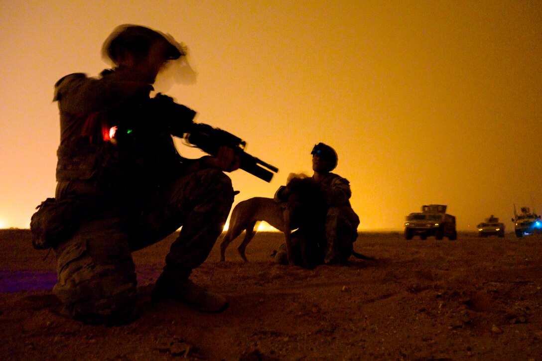 An airman kneels on the ground next to another airman holding a dog in front of parked military vehicles.