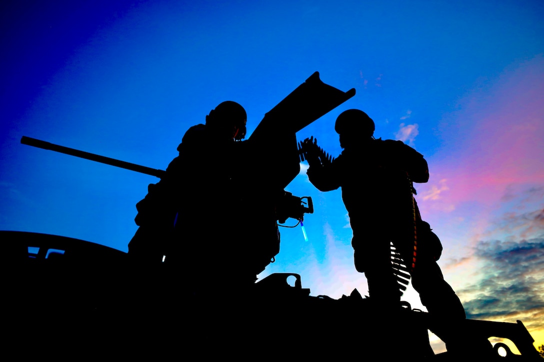 Two soldiers, shown in silhouette, stand atop a vehicle and load ammunition.
