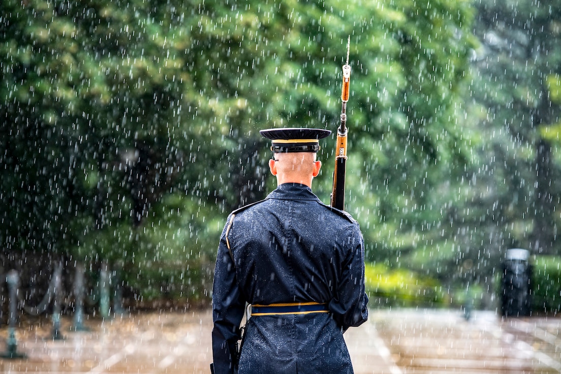 A soldier, seen from behind, stands and holds a rifle outside as rain pours down.
