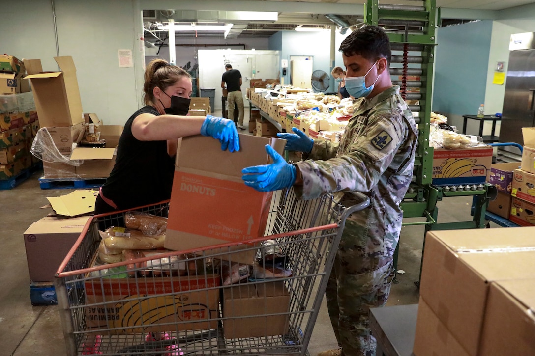Soldiers wearing face masks lift a cardboard box in an industrial kitchen-type room.