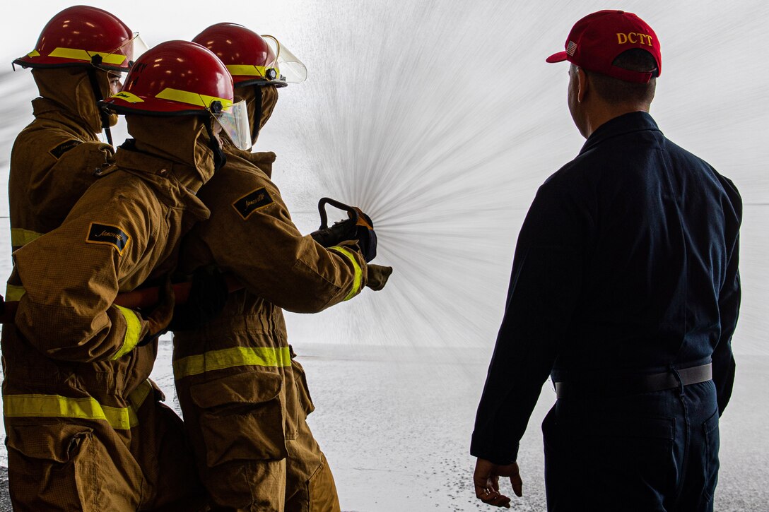 Three sailors in firefighting gear practice handling a hose while another man watches.