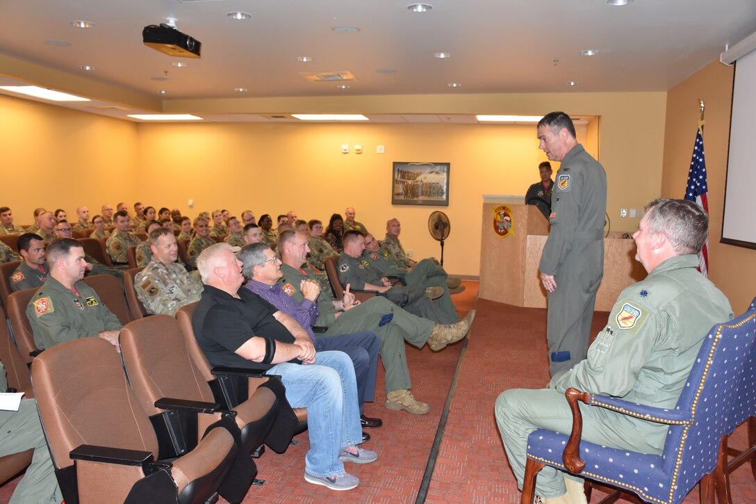A man speaks, and another man sits in a chair on a dais facing a bored crowd, but from a slightly different angle.
