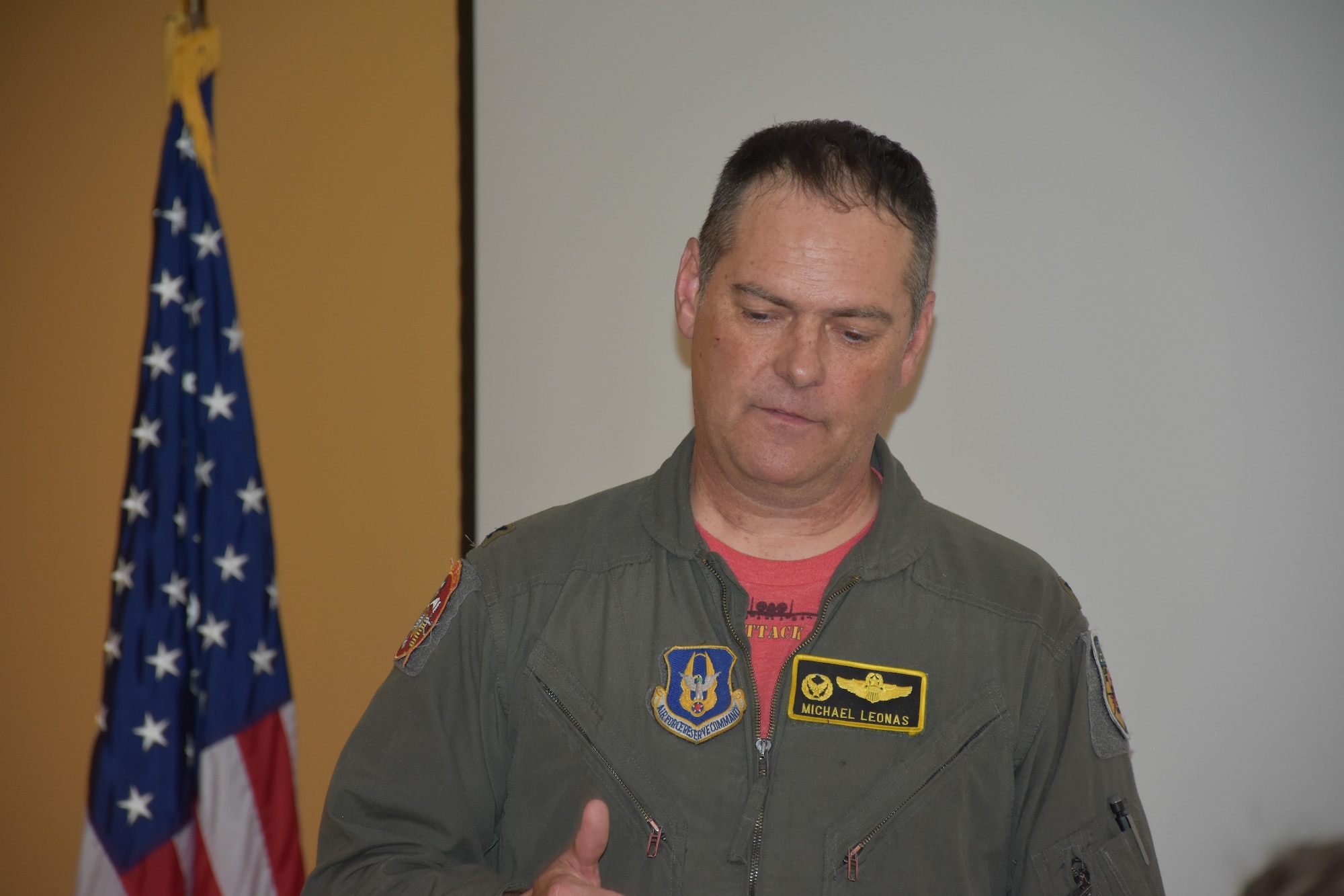 A man in a green flight suit with a red undershirt stands next to an American flag.