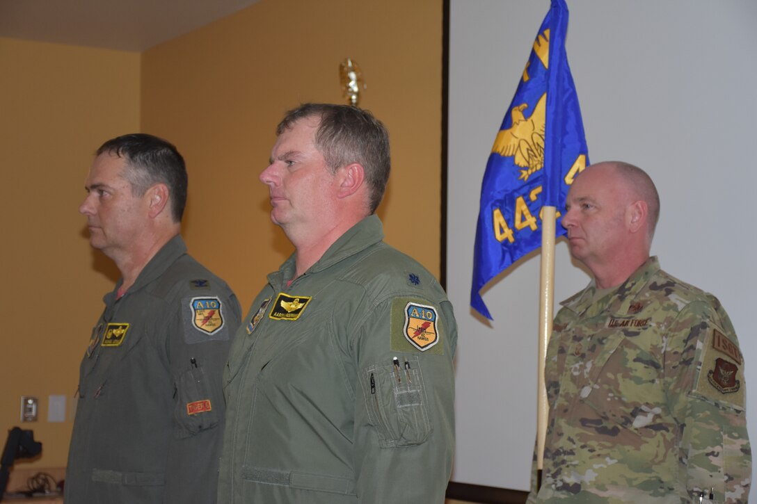 Two men in green flight suits stand in front of a man in camouflage who is holding a guidon.