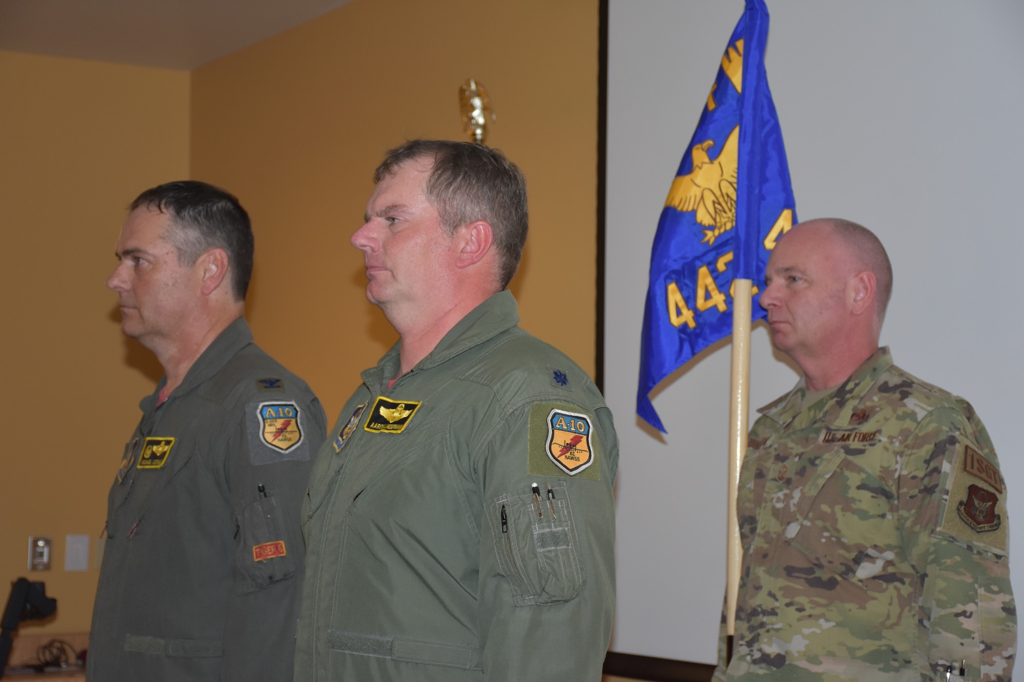 Two men in green flight suits stand in front of a man in camouflage who is holding a guidon.