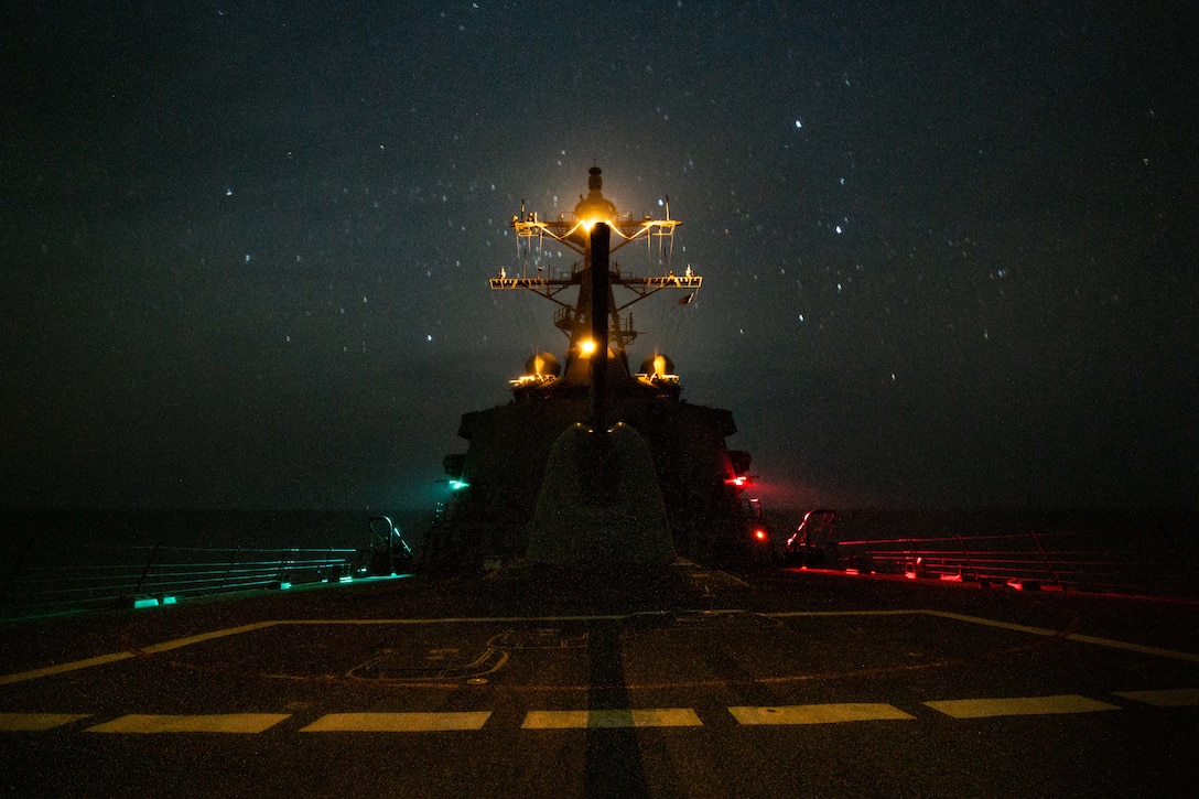 A ship sails under a starry night sky illuminated by white, green and red lights.