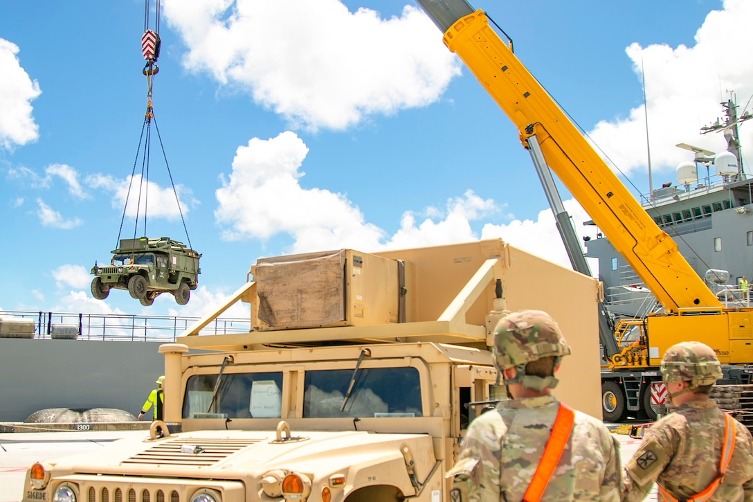 Two soliders watch a military vehicle being lifted by a yellow crane while standing near another vehicle.