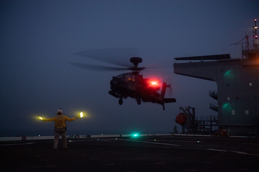 A sailor signals to a helicopter as it prepares to land on a ship at sea illuminated by colorful lights.