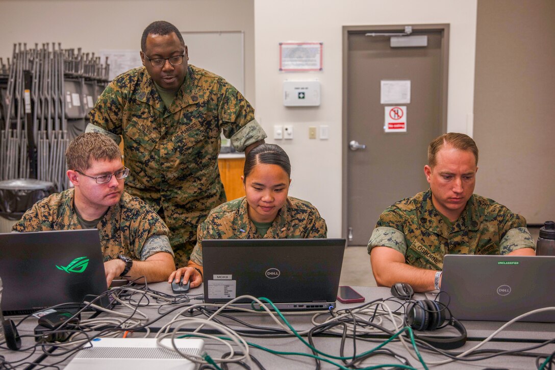 Three Marines work on laptops as one looks on.