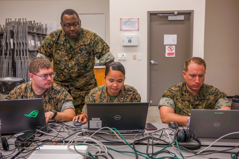 Three Marines work on laptops as one looks on.