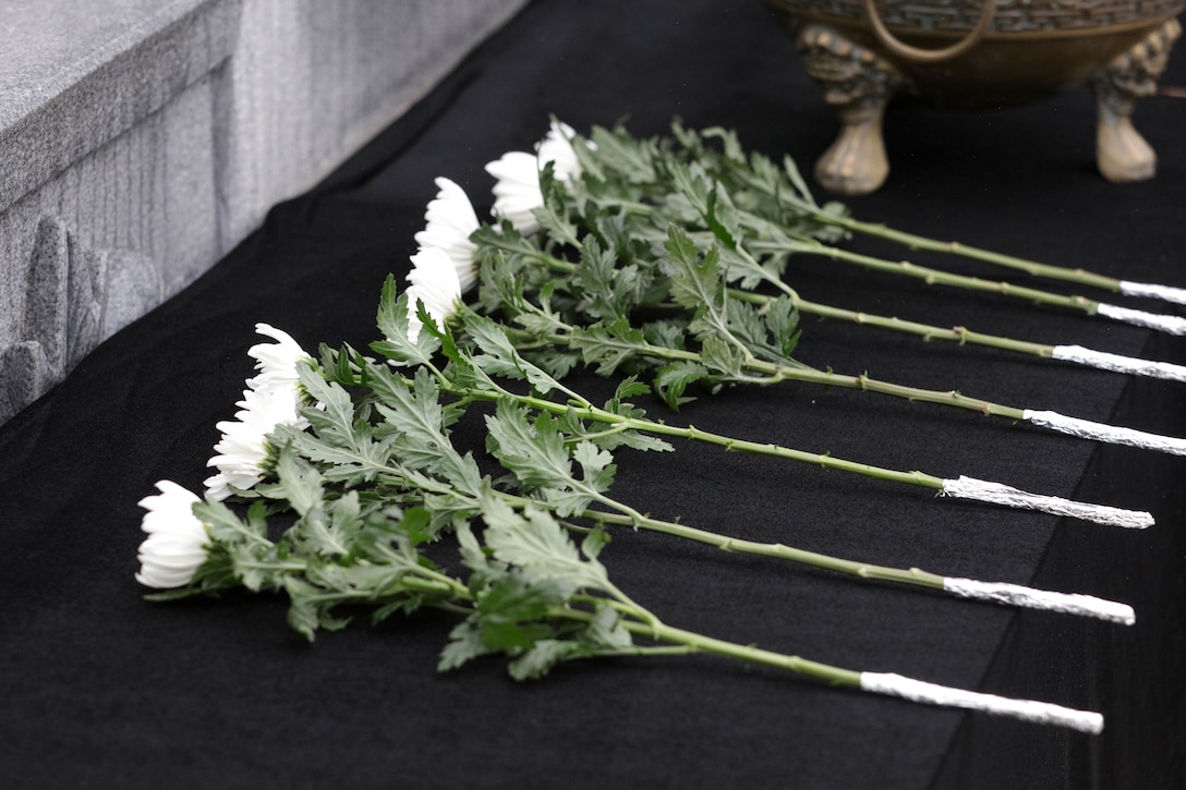Seven white flowers lay on a black cloth in front of the memorial stone.