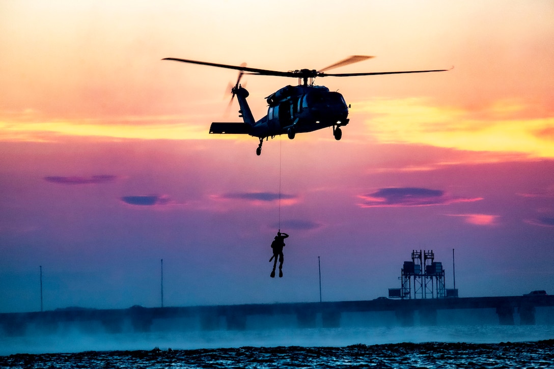 A sailor hangs onto a pulley as he dangles from a helicopter at twilight.