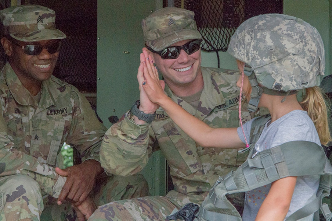 Two soldiers smile as they greet a young girl wearing a military helmet.