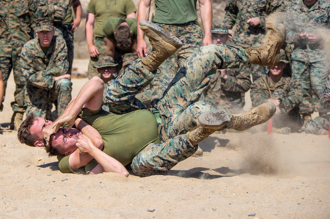 Two Marines grapple on sand as others watch.