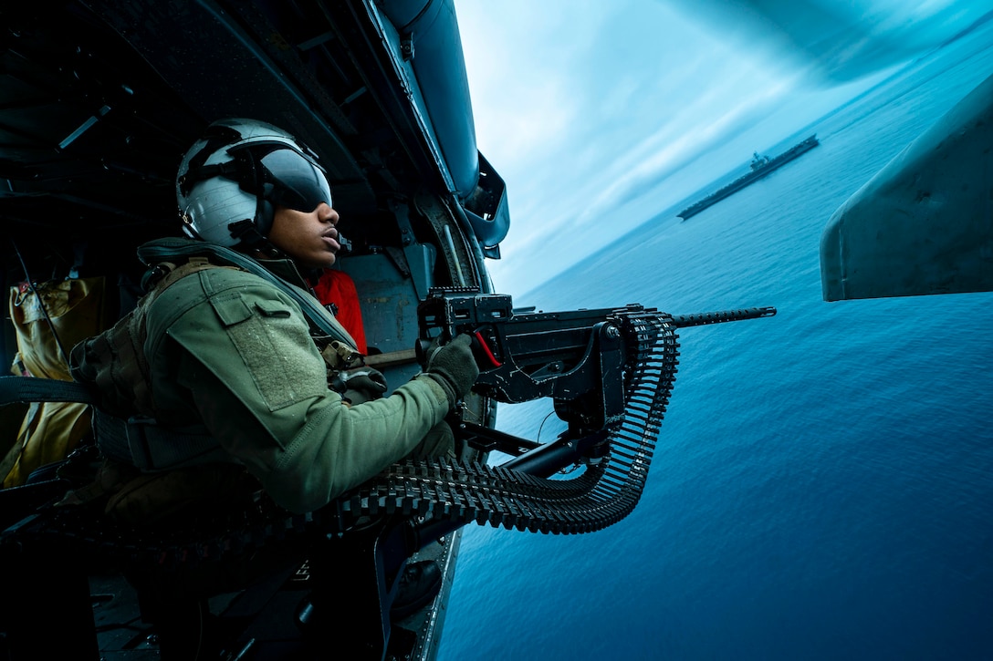 A sailor points a weapon out a helicopter door as it flies over water