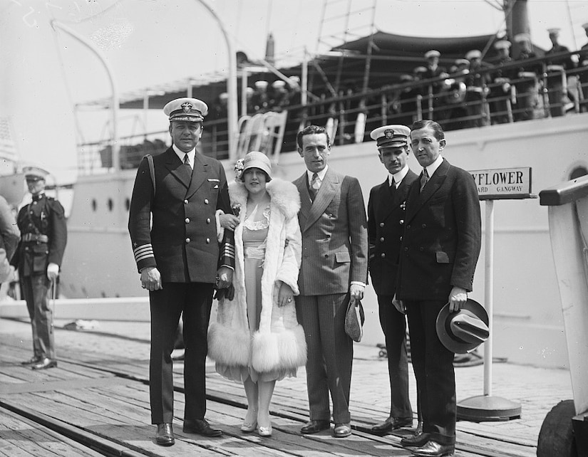 Five people in 1920s-style garb pose for a photo on the dock beside a yacht filled with sailors.