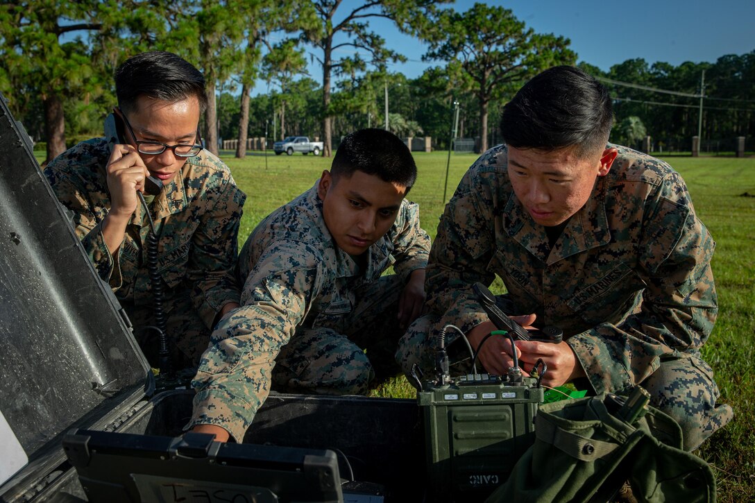 Three Marines crouch next to and test communications equipment in a field.