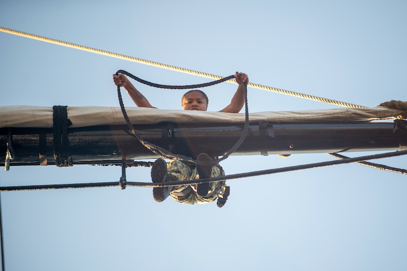 A sailor balancing on rope drapes another rope over the mast, as seen from below.