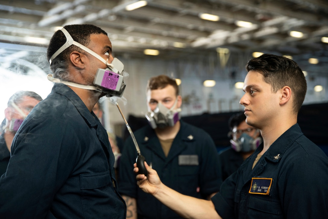 A sailor holds a chemical up to another sailor wearing a gas mask as fellow sailors observe.