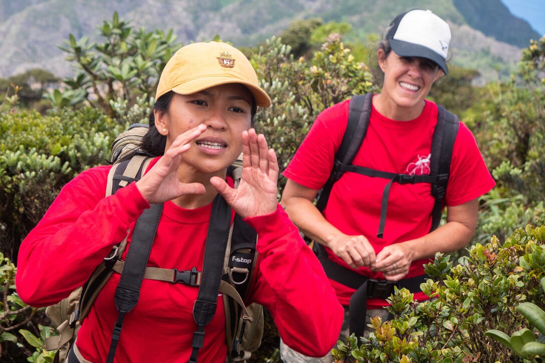 A female in a red shirt puts her hands up to her mouth to talk to people as another female in a red shirt looks on with mountains in the background.