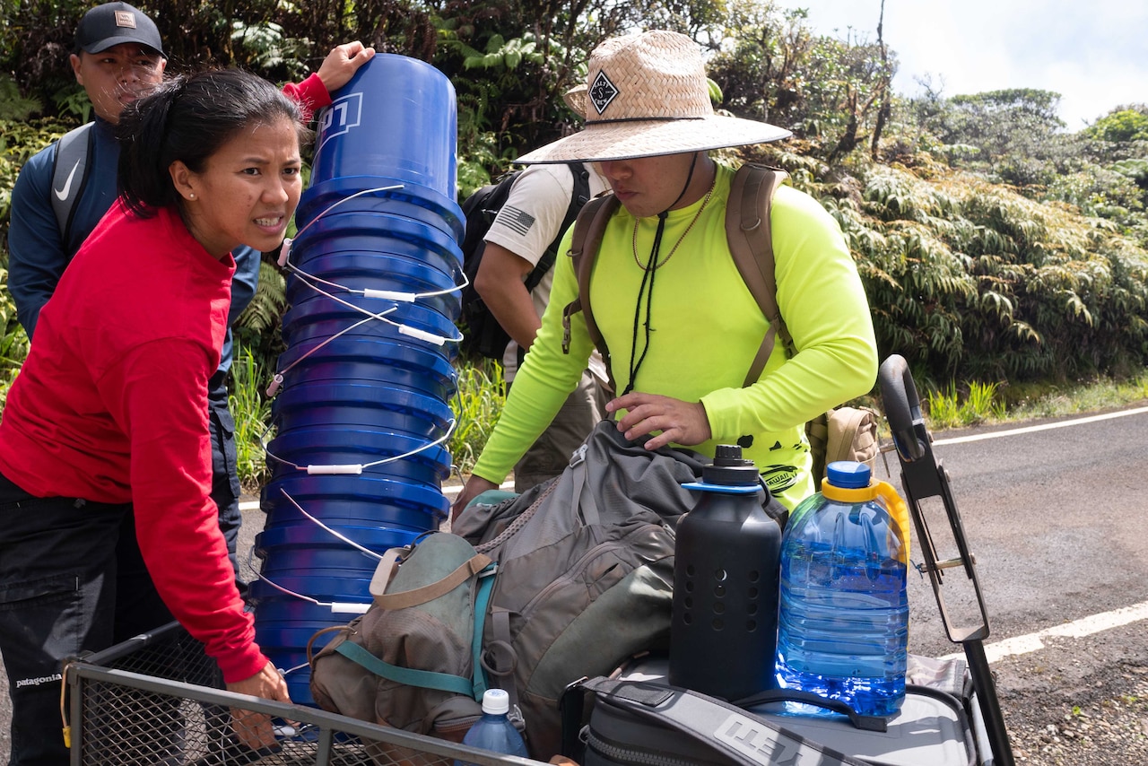 A female airman in a red shirt grabs a stack of blue buckets from a cart while an airman with a yellow shirt grabs a gray bag.