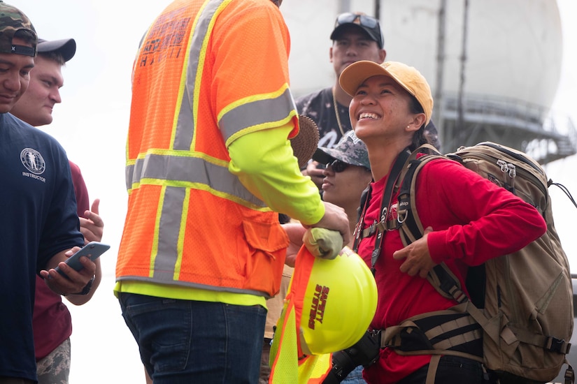 A female airman smiles at a man in a safety vest holding a yellow hard hat.