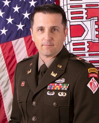 White male with brown hair, dressed in a brown uniform with medals and rank, stands in front of the red, white and blue American flag and the red and white Army Corps flag.