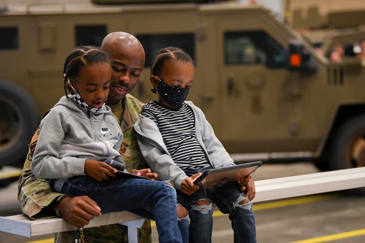 An airman stands behind two seated children looking at tablets.