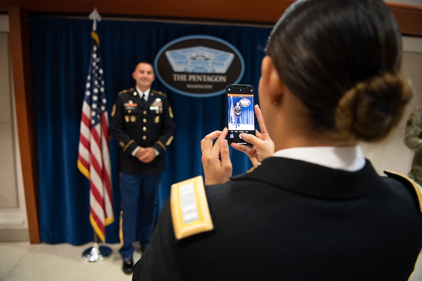 a woman takes a photo with her cell phone of a man in an army uniform.