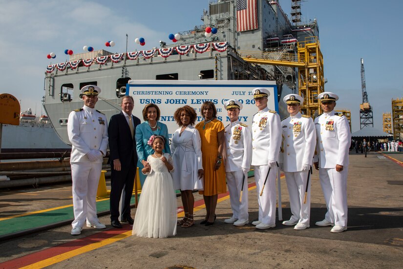 A group of Navy officers and civilians stand on a dock in front of a large ship.