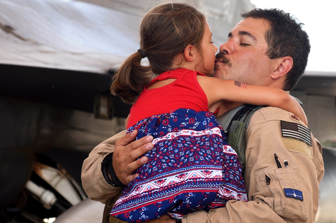 An airman hugs and kisses his daughter next to an aircraft.