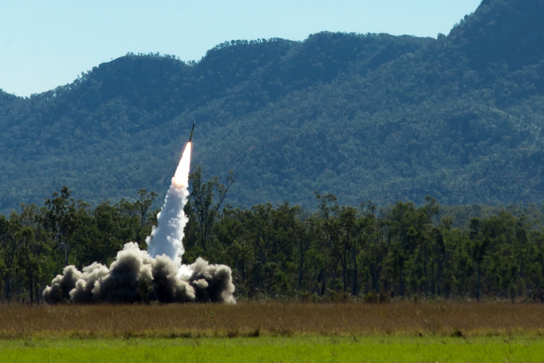 A missile launches in a field with hills in the background leaving smoke behind.