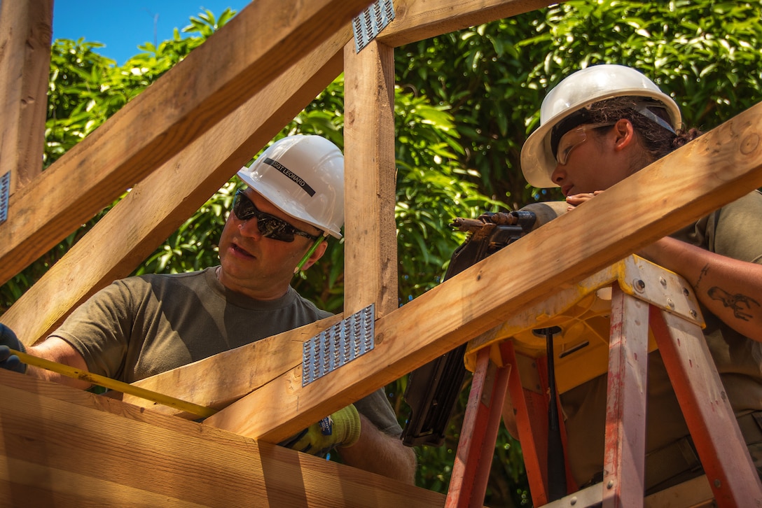 An airman uses a measuring tape while instructing another within a building frame.