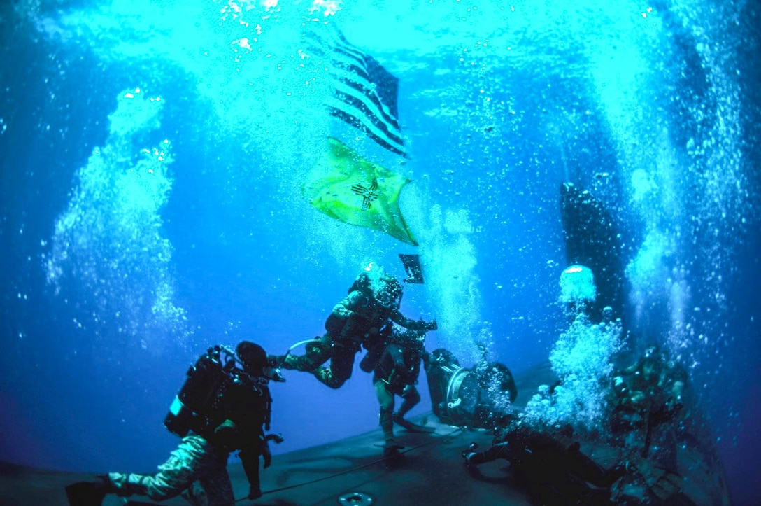 Divers swim by a sub underwater, where an American and a New Mexico flag also wave.