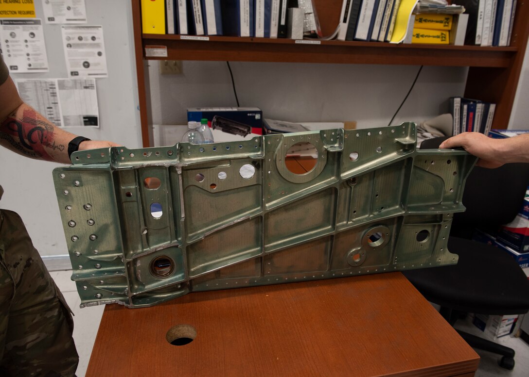 Two Airmen hold a damaged trunnion beam on a desk.