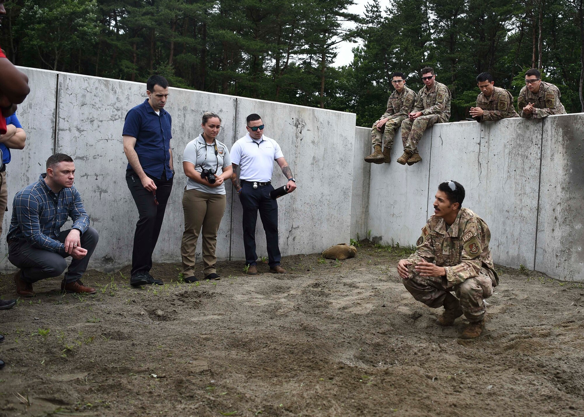 A military member squats in front of other military members in a roofless concrete box