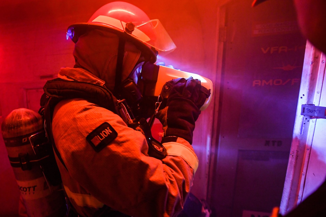 A sailor dressed in firefighting gear looks through a device.