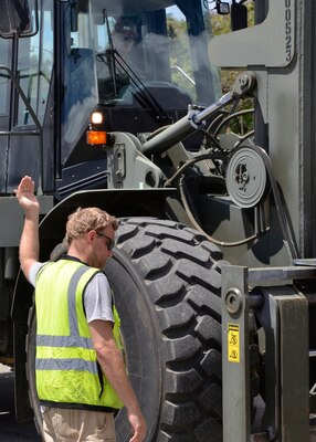 A man stand next to a large green excavator giving directions to the driver by the use of his hands
