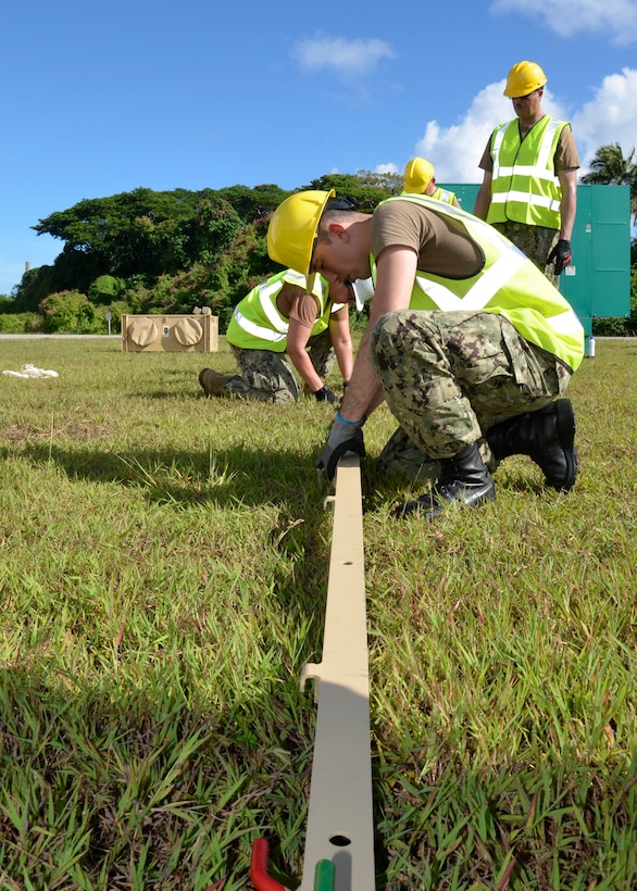 A man in a hard hat and safety vest kneels down to work on a long post that is part of the tent he is putting together