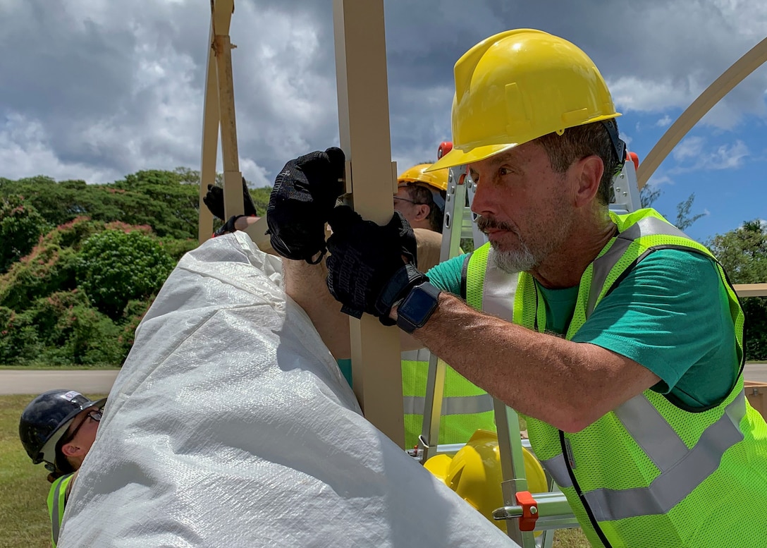 A man wearing a hard hat looks closely at two upright supports of a tent as he places a pin into the them to connect them to each other.