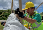 A man wearing a hard hat looks closely at two upright supports of a tent as he places a pin into the them to connect them to each other.