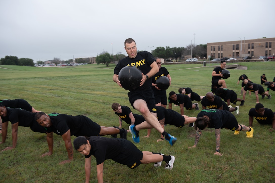 A soldier runs with an exercise ball while others do pushups in a large field.