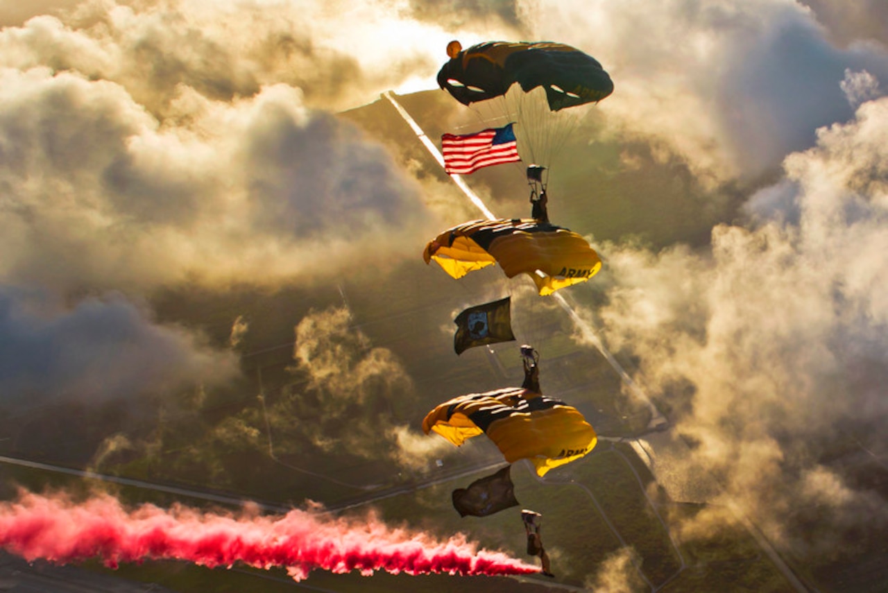 Three service members freefall wearing parachutes near clouds of red smoke.