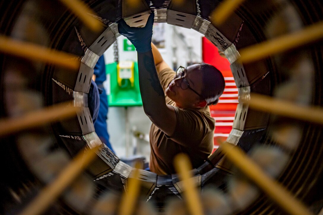 A sailor is framed by the circular opening of an afterburner she is working on.