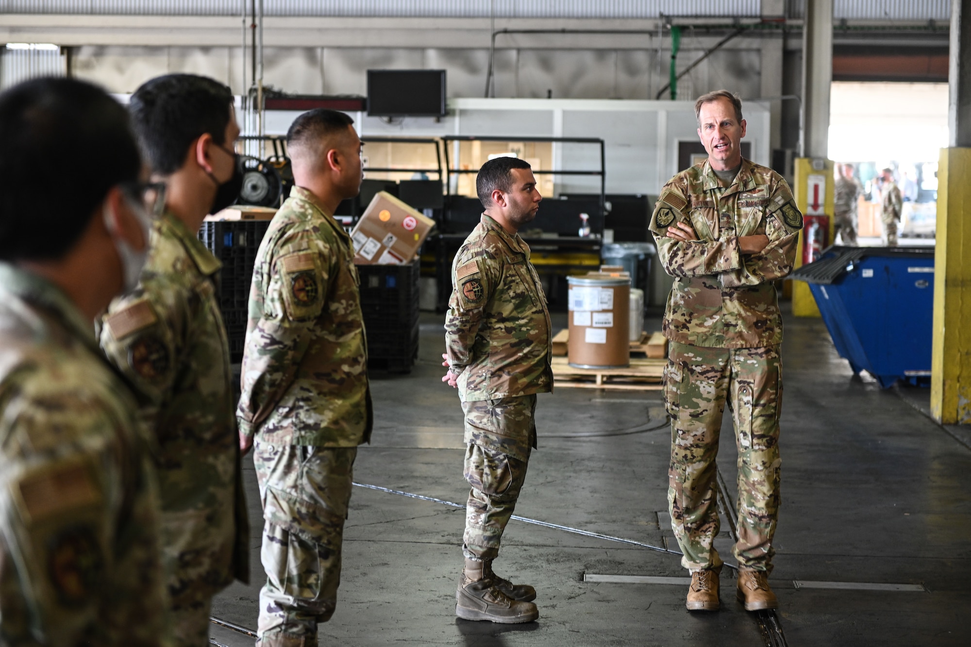 A man in a military uniform speaks to others in formation inside of a warehouse