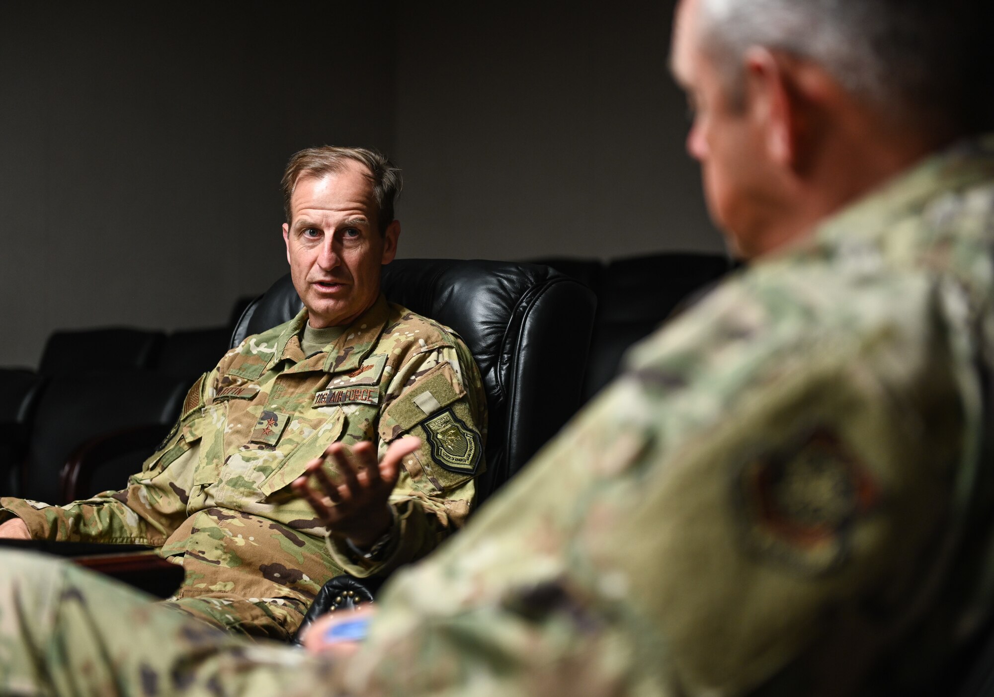 A man in a military uniform speaks to another inside of a dimly-lit conference room