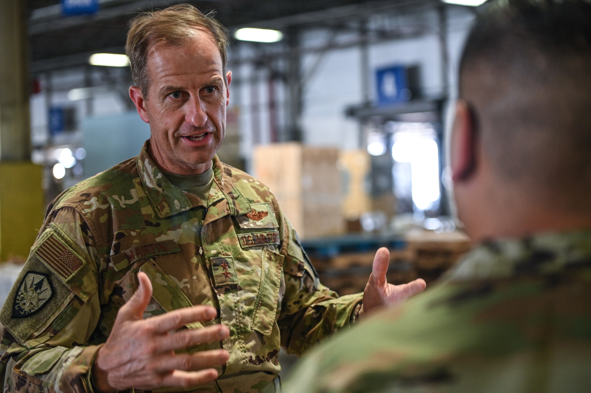 A man in a military uniform speaks to another inside of a warehouse.