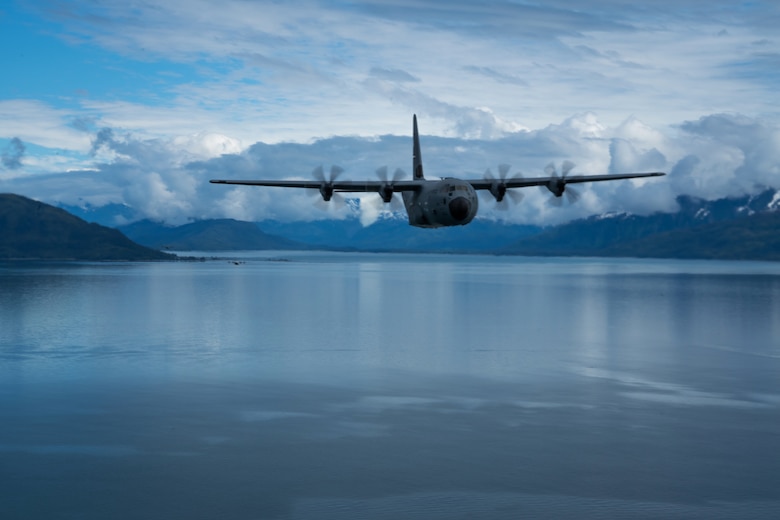 An aircraft flies over a body of water near snowy mountains.