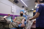 Military service member is reclined inside of the KNRC bus donating blood. A KNRC nurse stands to the side drawing donator's blood.